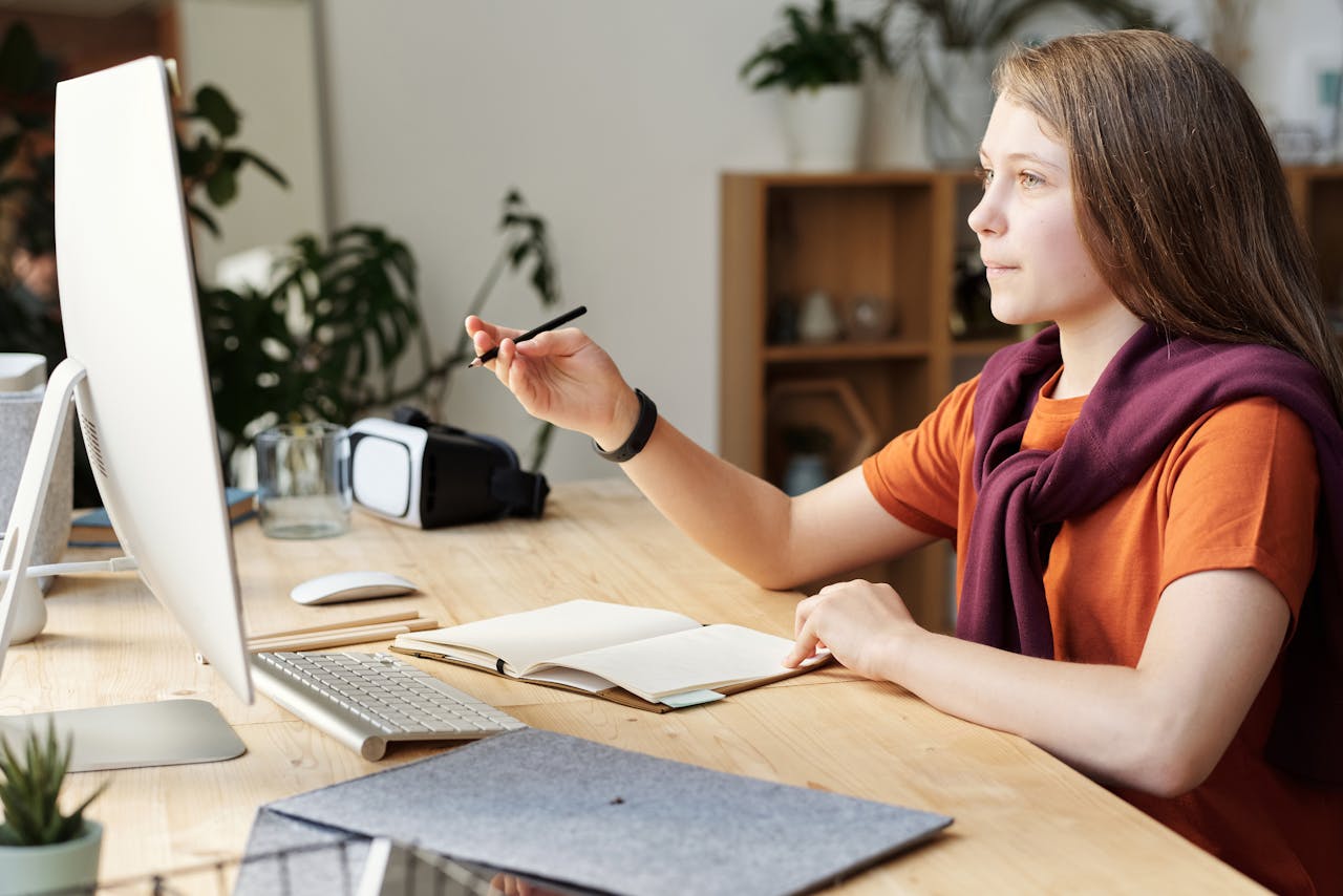Teen girl in home office studying online using a computer and writing in a notebook.
